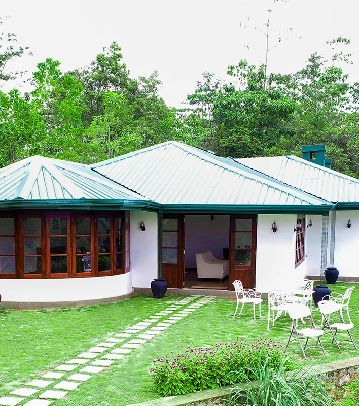 A white bungalow with a green roof surrounded by lush greenary